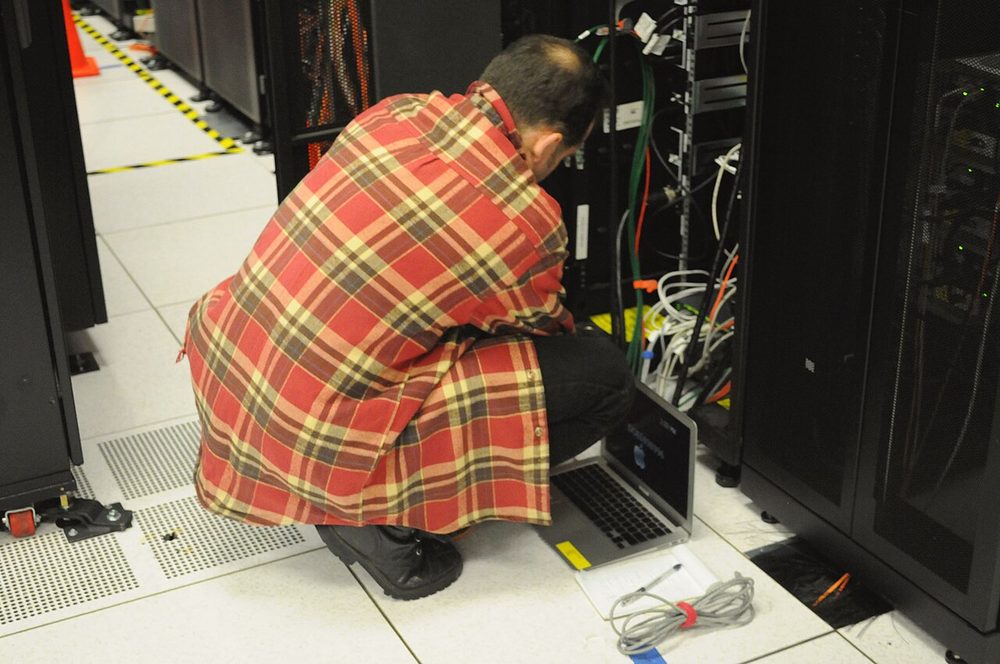 A technician checking a server rack with a laptop at a computing centre